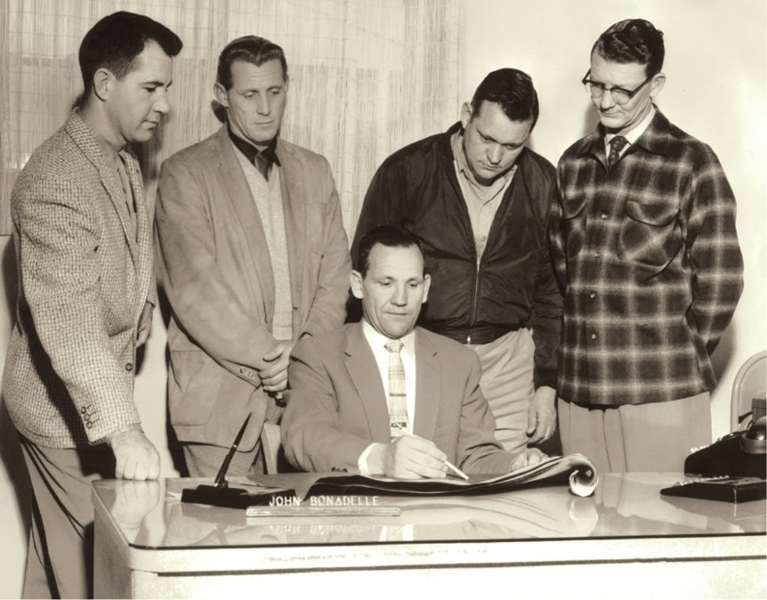 A team gathers around Mr. Bonadelle's desk.